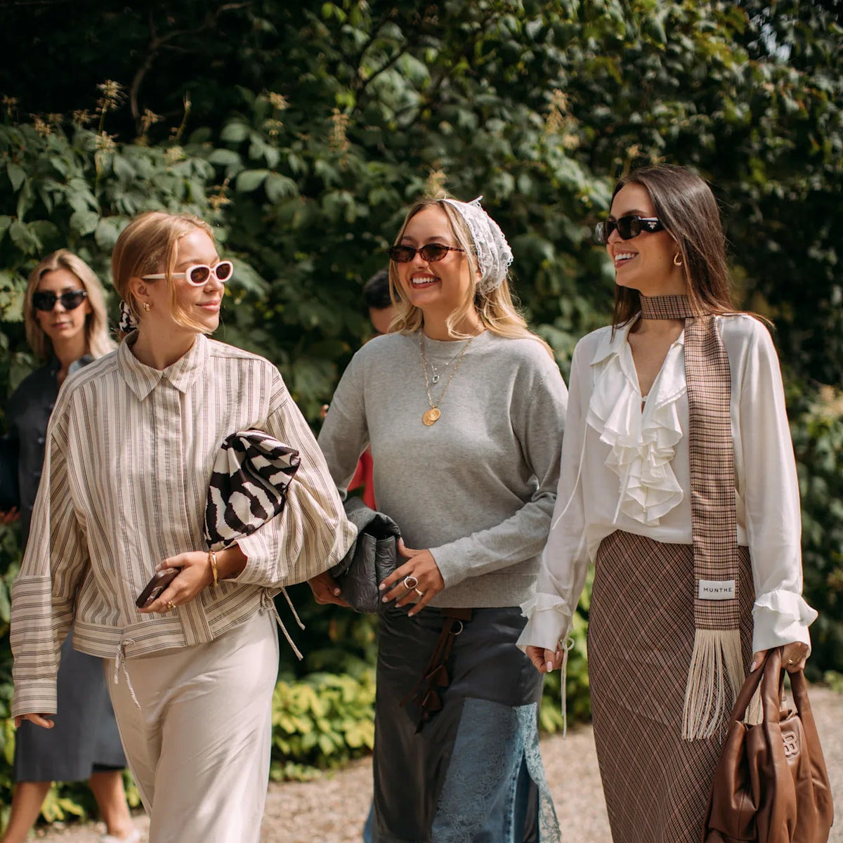 Three women walking outdoors on a gravel path surrounded by green foliage, showcasing stylish fashion looks with neutral tones.